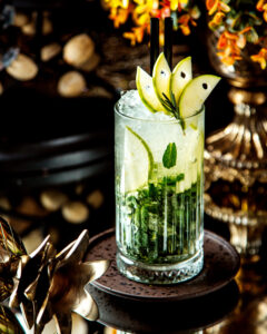 A crystal glass filled with a mojito, garnished with fresh mint leaves and slices of green apple arranged elegantly, creating a refreshing and stylish presentation.