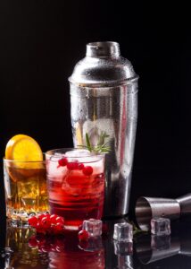 Two cocktails, a whiskey with orange slices and a vodka cranberry with red berries, placed next to a metal cocktail shaker on a reflective surface with ice cubes.