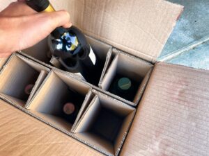 A person placing a bottle of wine into a cardboard delivery box with compartments holding multiple bottles.