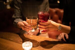 Two people clinking glasses, one holding a whiskey and the other a red cocktail, in a cozy bar setting with warm lighting.