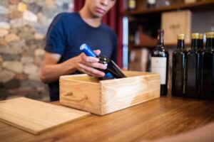 A worker carefully packing a bottle of wine into a wooden crate, preparing it for delivery alongside other bottles on the counter.