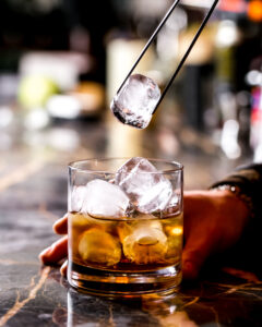 A bartender using tongs to place ice cubes into a glass of whiskey, set on a marble bar counter, creating a chilled and refined drink.