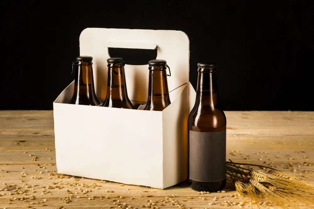 A six-pack of brown glass beer bottles in a white carton box with one bottle placed next to it, on a wooden table with wheat ears and grains.