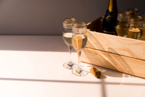 Two champagne flutes filled with sparkling wine placed on a sunlit white table, next to a wooden crate with more bottles and a cork.