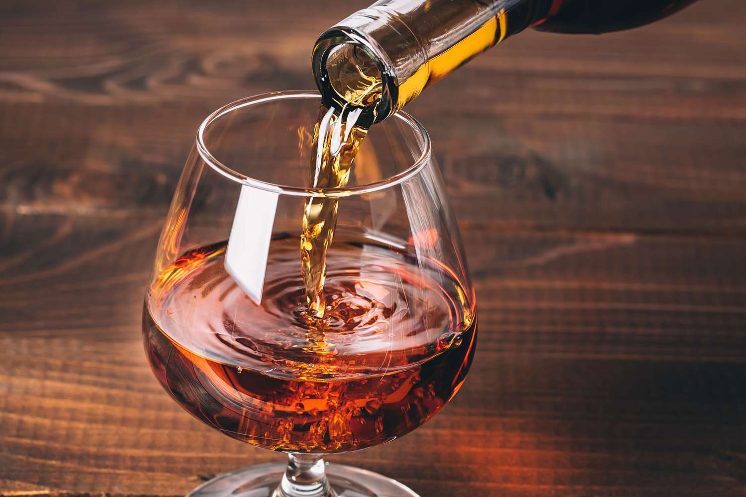 A glass of cognac being poured, showcasing the rich amber color of the liquor as it swirls in the glass, set against a rustic wooden background.