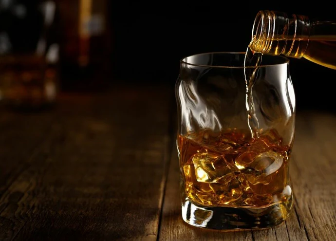 A glass of whiskey with ice cubes being poured from a bottle, set on a rustic wooden table, showcasing the rich amber color of the liquor.