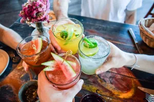 A group of people toasting with colorful cocktails, featuring watermelon, lime, and jalapeño garnishes, at a table decorated with a vase of flowers.