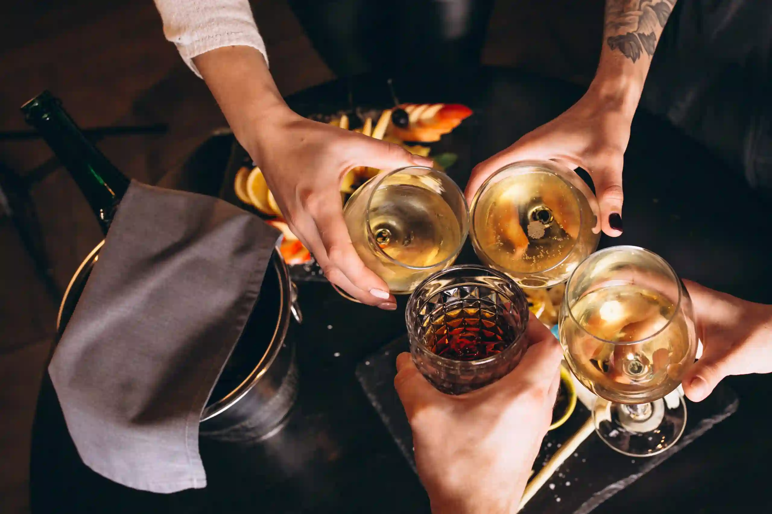 Group of people clinking glasses with white wine and cocktails at a cozy gathering, with a table of appetizers in the background.