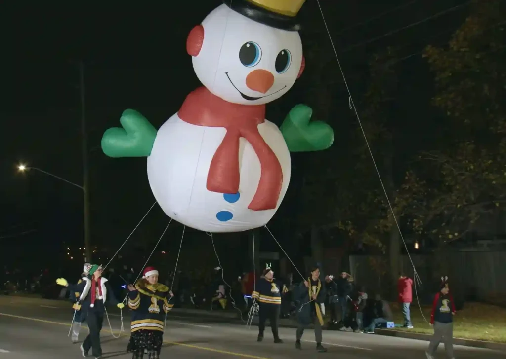 Large inflatable snowman float at the Brampton Christmas Parade, surrounded by people guiding it down the street at night.