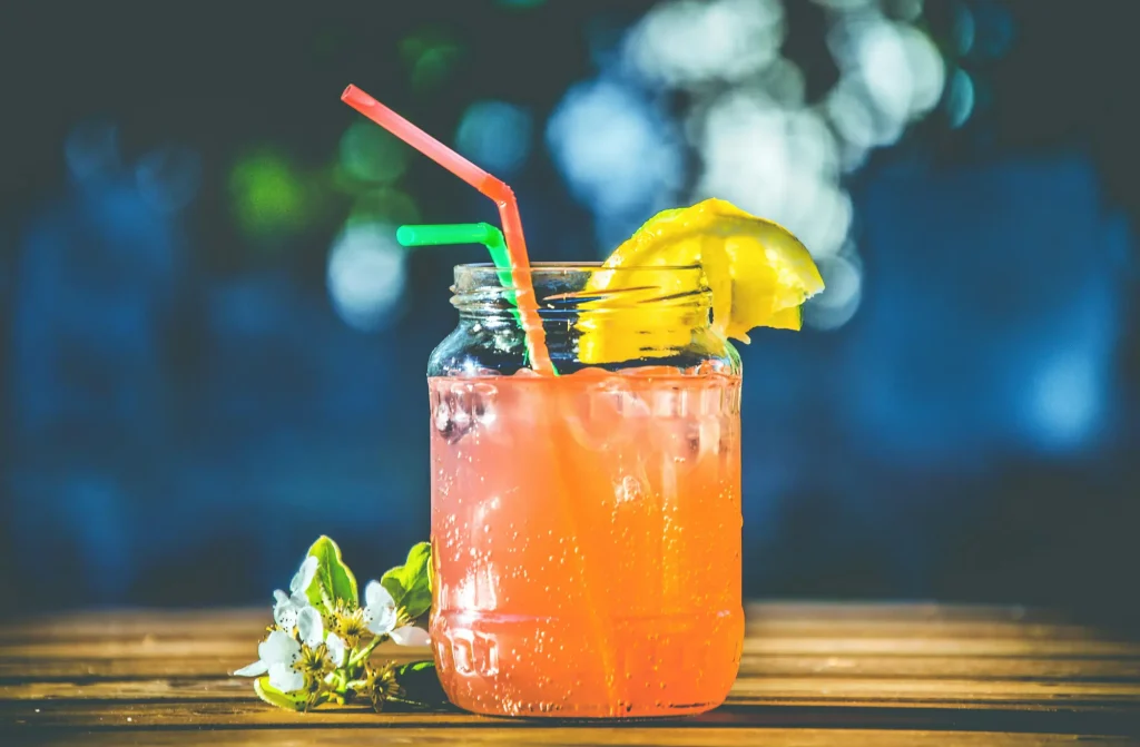 A vibrant orange drink in a mason jar with a red and green straw, garnished with a lemon slice and accompanied by small white flowers on a wooden table.