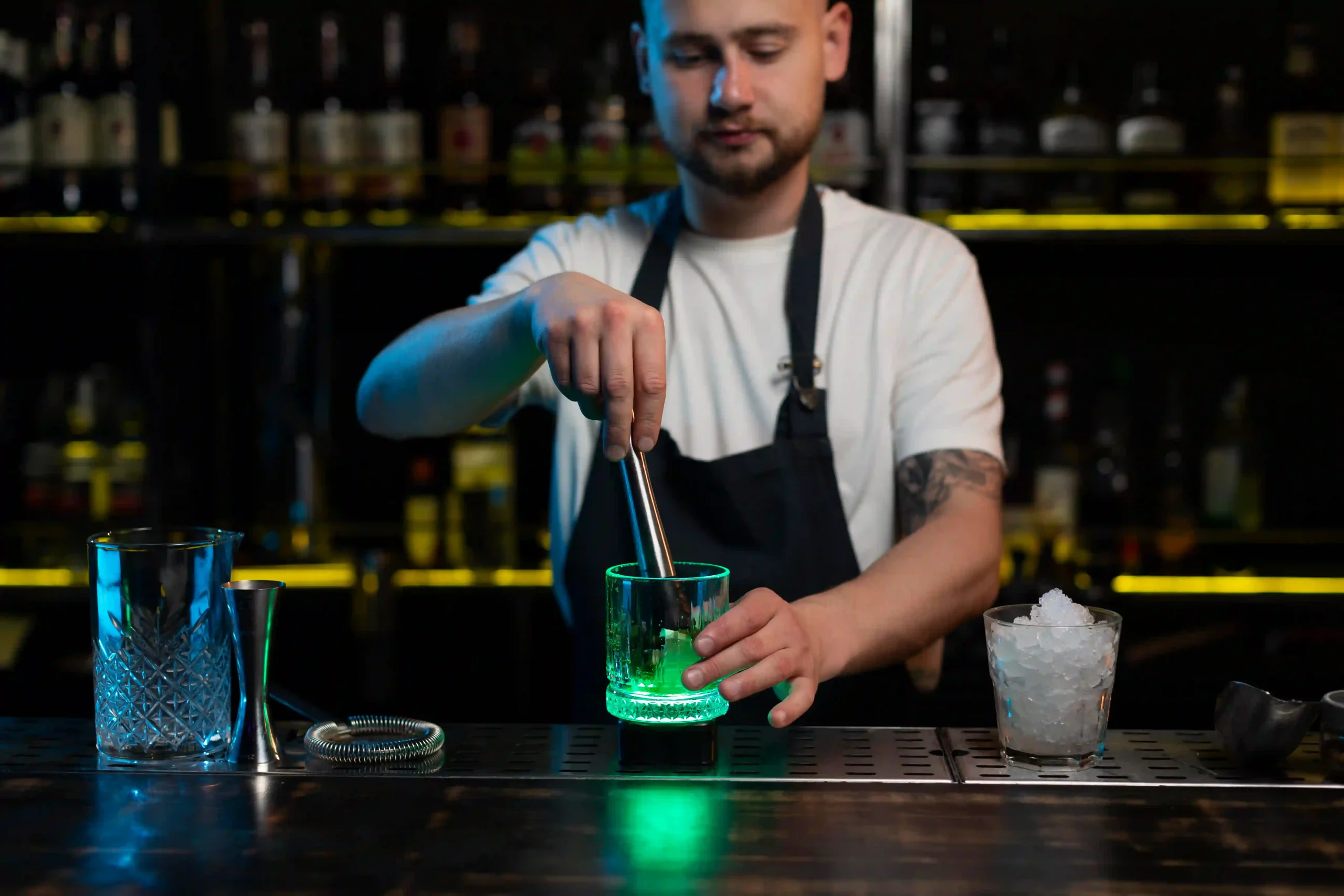 Bartender preparing a cocktail at a bar counter with a green-lit glass and bartending tools.