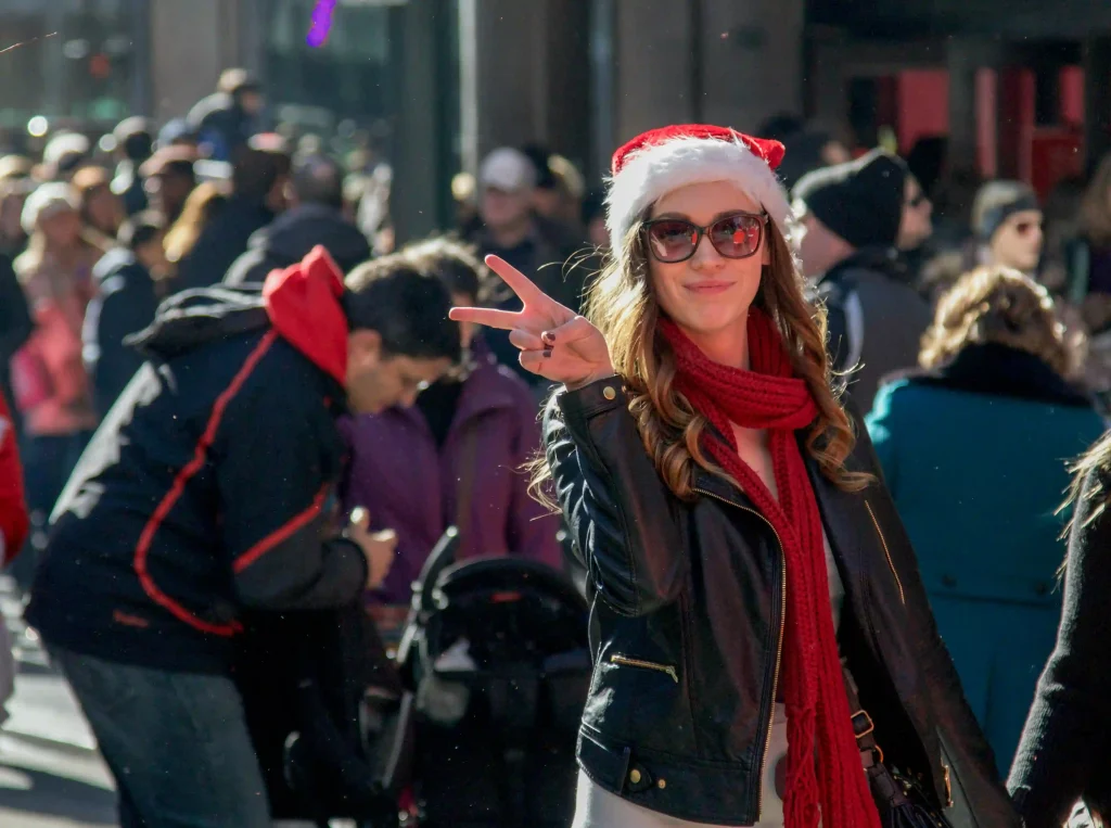 Smiling woman wearing a Santa hat and red scarf, making a peace sign at a Christmas parade with a crowd in the background.