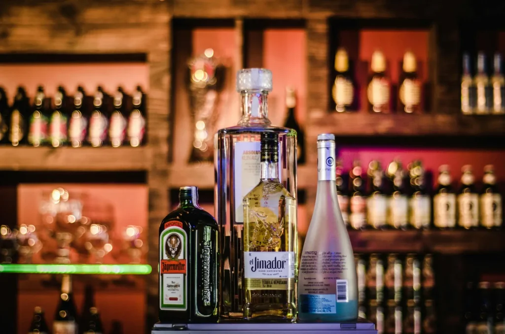 A selection of liquor bottles, including Jägermeister, El Jimador tequila, and Hpnotiq, displayed on a bar counter with a blurred background of additional bottles on shelves.