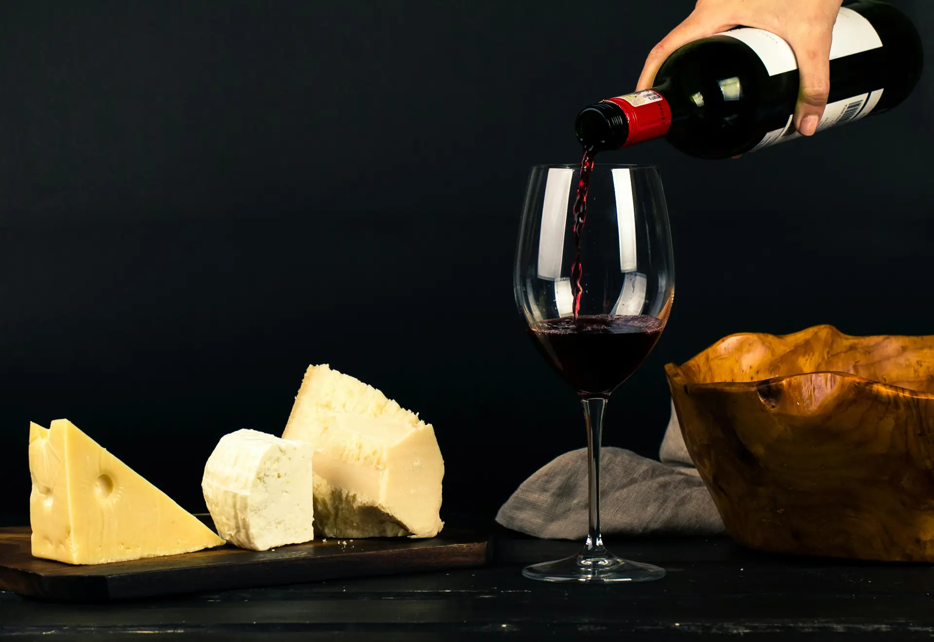 Red wine being poured into a glass alongside a wooden platter of assorted cheeses.